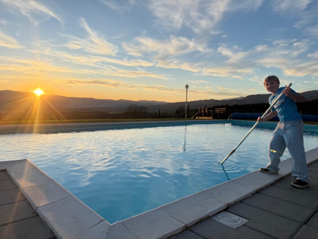 Ein Junge reinigt mit einem langen Besen einen Swimmingpool im Sonnenuntergang während des Trainingslagers in der Toskana.